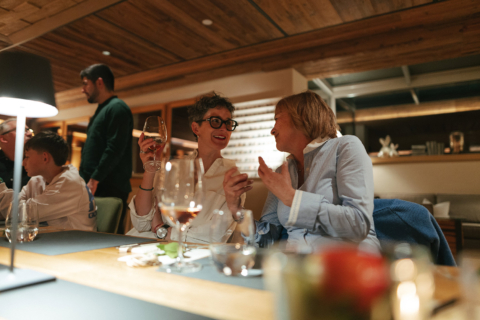 Zwei Frauen lachen und unterhalten sich am Community Table bei einem Glas Wein in gemütlicher Atmosphäre