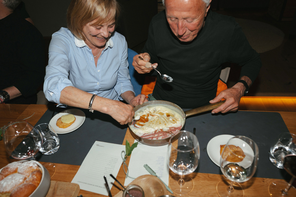 Zwei Gäste teilen sich am Community Table eine Pfanne mit geschmolzenem Käse und genießen gemeinsam den Moment